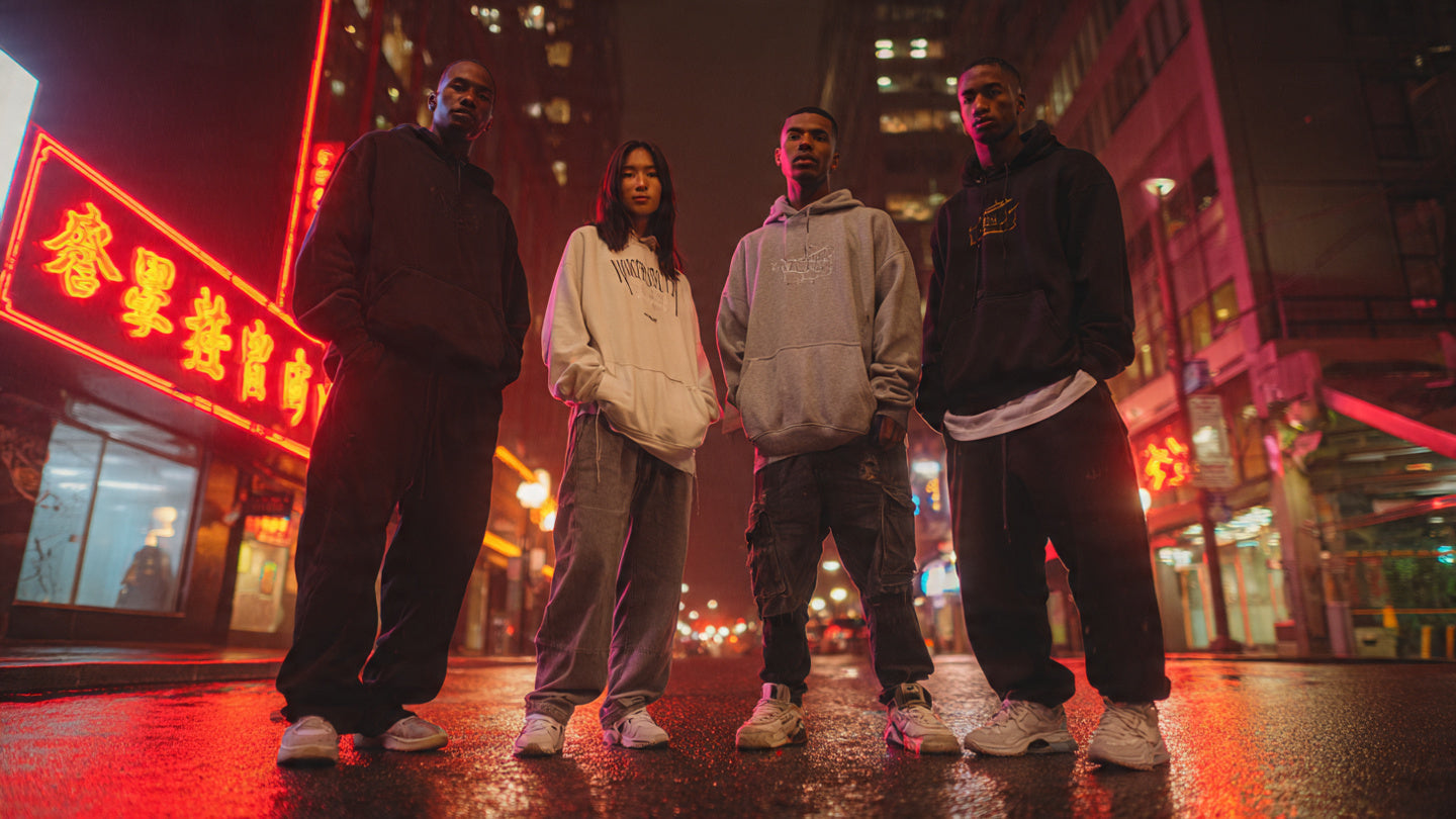 Group of young adults in streetwear hoodies standing on a neon-lit city street at night.